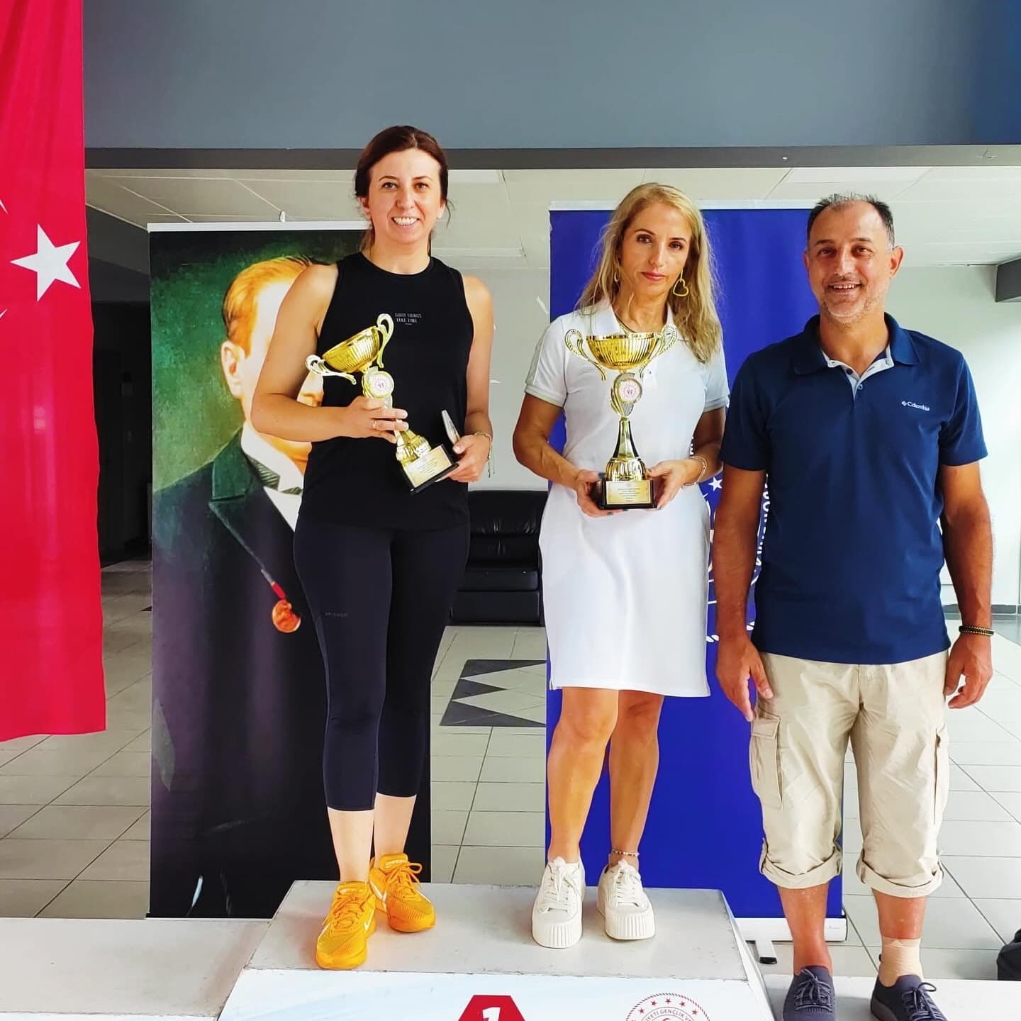 Kadınlar turnuvası ödül töreninde kupalarını tutan iki sporcu ve turnuva görevlisi