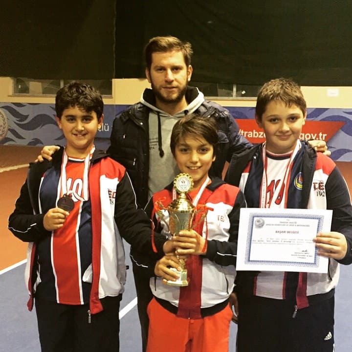 Salon kortunda kupa ve başarı belgeleriyle poz veren üç erkek sporcu ve antrenörleri