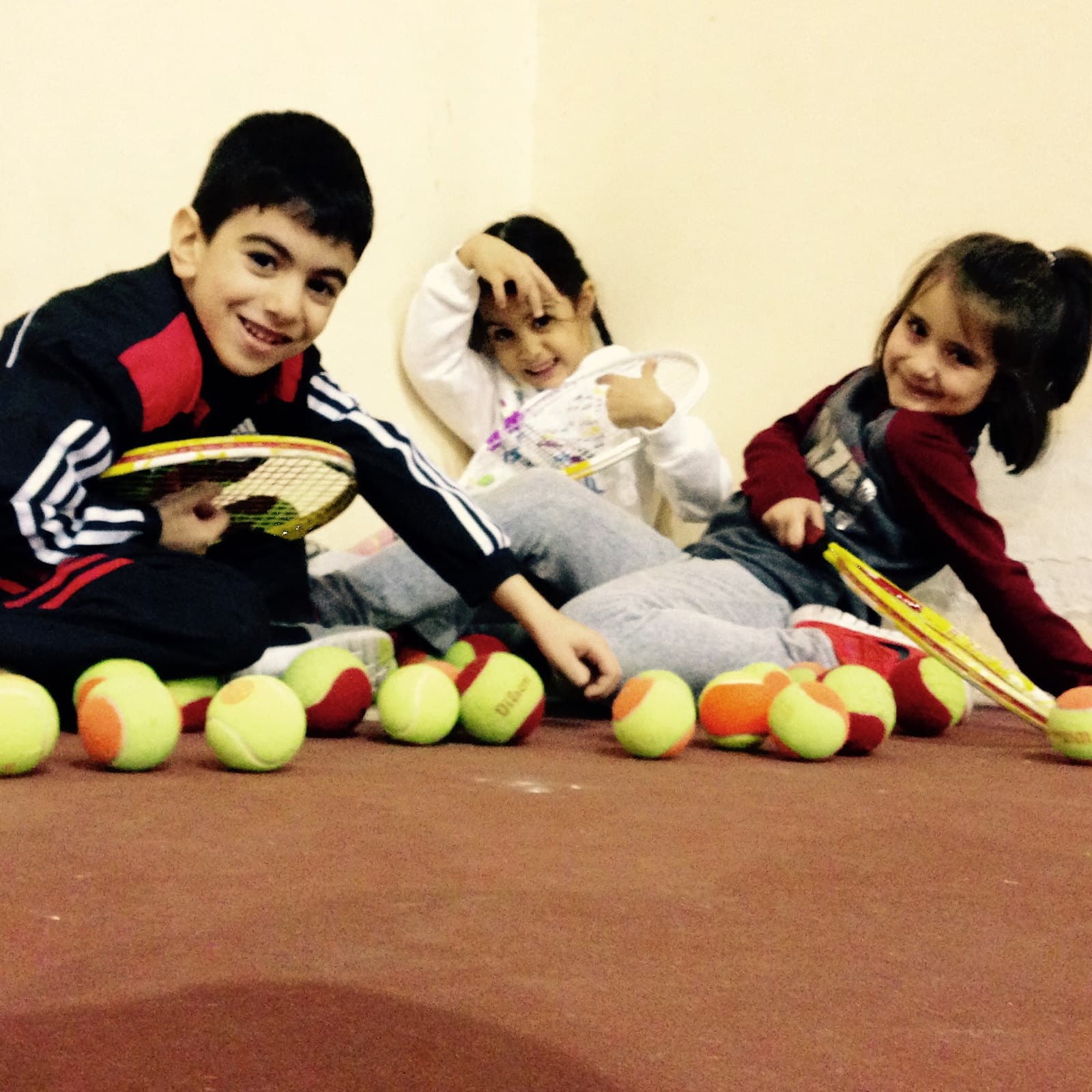 Kapalı kortta tenis toplarının arasında raketleriyle oynayan üç küçük çocuk